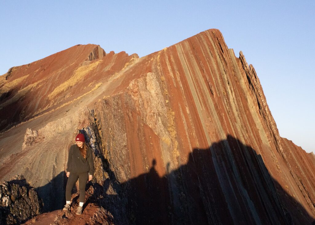 Megan standing at the viewpoint in front of the multi-coloured Pallay Punchu Mountain at golden hour.