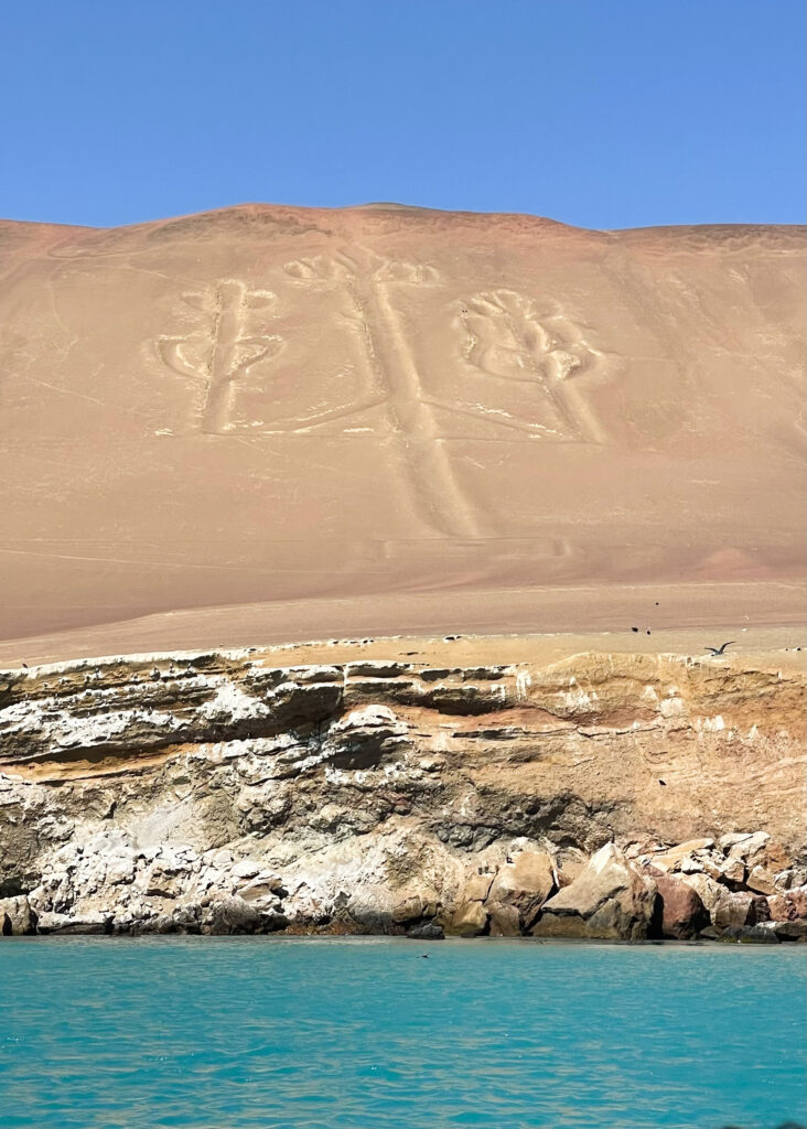 The Paracas Candelabra. A trident-like shape etched into the sand on the side of a cliff, viewed from the a boat in the sea. 