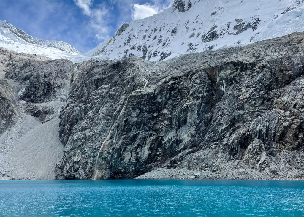 Landscape photo of Laguna 69. The water is bright blue and behind there are snow-capped mountains with a small waterfall feeding into the lake. 