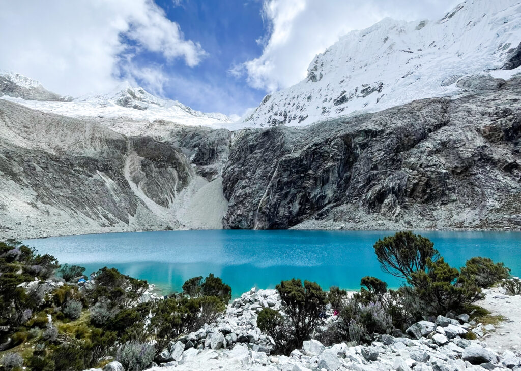Landscape photo of Laguna 69. The water is bright blue and behind there are snow-capped mountains with a small waterfall feeding into the lake. 