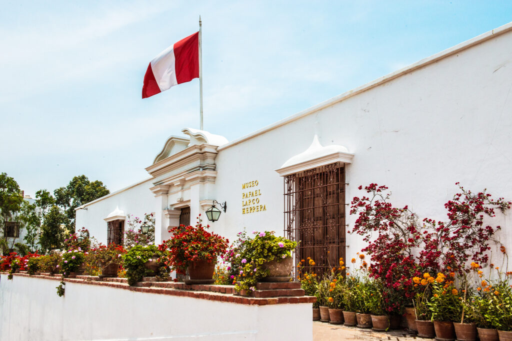 The outside of the Larco Museum. A beautiful white building with flowers at the front and a Peru flag above. 