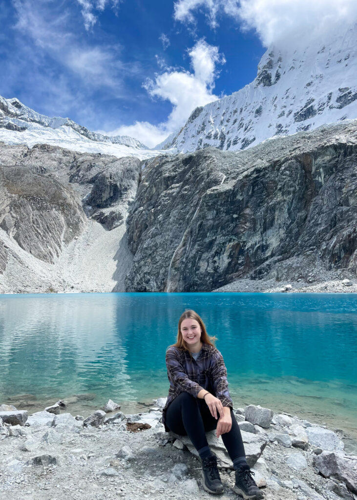 Megan sat in front of Laguna 69. The water is bright blue with snow-capped mountains behind and a small waterfall feeding into the lake