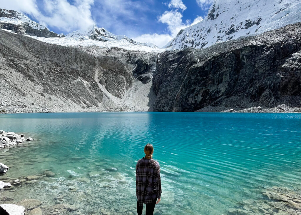 Megan stood at the shore of Laguna 69. The water is bright blue and behind there are snow-capped mountains with a small waterfall feeding into the lake. 