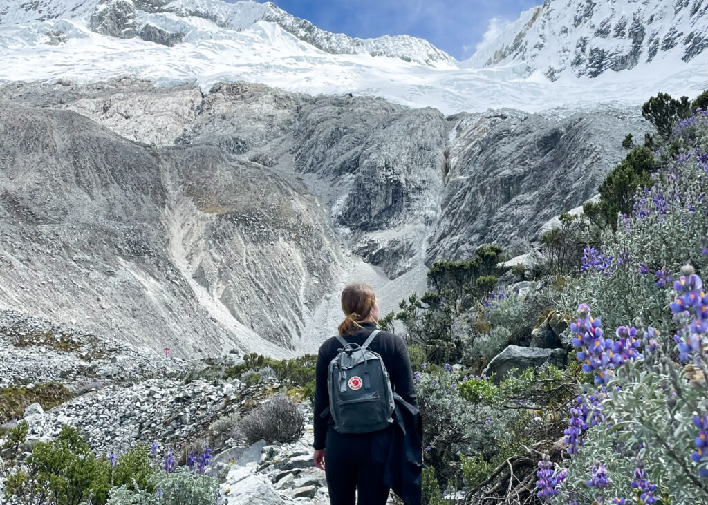 Megan on the Laguna 69 hike with snow-capped mountains in the background and purple flowers in the foreground. 